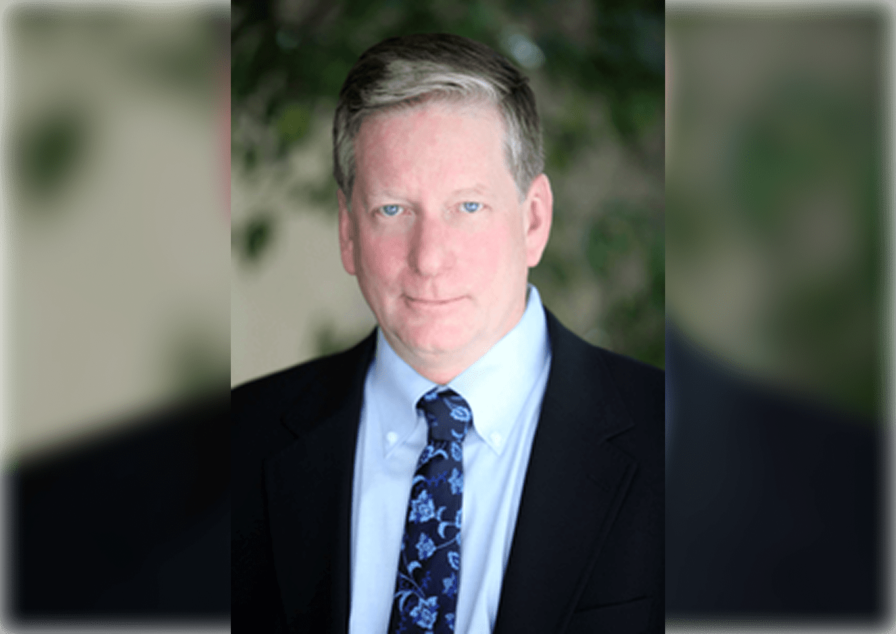 A man in a suit and blue tie stands against a blurred green background, looking at the camera.